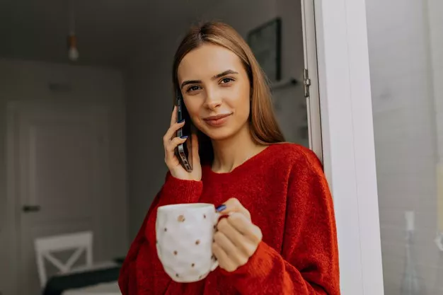 estilizando uma mulher espetacular com cabelo castanho claro vestindo uma blusa vermelha usando smartphone e tomando café em casa pela manha